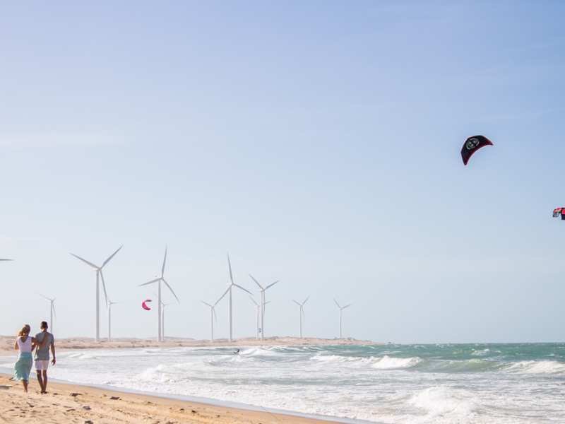Pessoa caminhando pela praia de Icaraizinho carregando prancha de windsurf e vela com turbinas eólicas ao longe