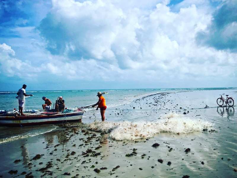 Pescadores na praia de Icaraizinho trabalhando com redes, criança ao lado com bicicleta e turbinas eólicas no horizonte