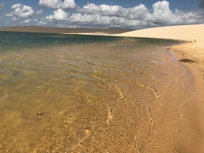 Mulher caminhando na areia próxima a uma lagoa dos Lençóis Cearenses em Caetanos, reflexo da duna na água